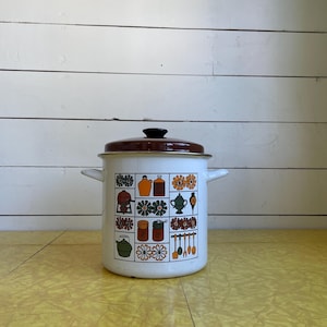 May include: White enamel cooking pot with a brown lid and black handle. The pot features a colorful retro design of kitchen-themed illustrations. The pot has two side handles and is set against a white wooden background and a yellow surface.