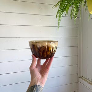 May include: A handmade ceramic bowl with a brown and tan glaze. The bowl has a ribbed exterior and a rounded shape. The interior is a lighter tan color, and the exterior has a darker brown glaze that drips down the sides.