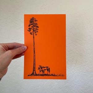 May include: A rectangular print featuring a vibrant orange background with a black ink illustration. The artwork depicts a tall, slender pine tree standing next to a mare and her foal grazing in a grassy field at the base of the tree.