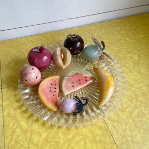 May include: A clear, decorative glass plate holds a collection of carved stone fruit. The assortment includes a red apple, a peach, a watermelon slice, a banana, and other fruits in various colors. The plate sits on a yellow surface.