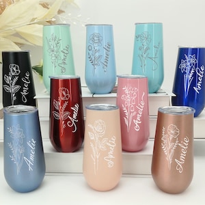 Könnte beinhalten: Eine Sammlung von Sektgläsern ohne Stiel in verschiedenen Farben, darunter Schwarz, Blau, Rot, Rosa und Gold. Jedes Glas hat ein Blumendesign und den Namen "Amelie" in Schreibschrift. Die Gläser werden auf einer weißen Oberfläche präsentiert.