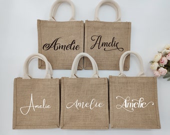 May include: Five small brown burlap tote bags with white handles. The bags have the name "Amelie" written on them in a cursive font. Some bags have black lettering and some have white lettering.
