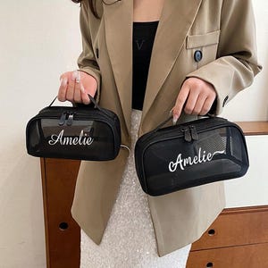 Könnte beinhalten: Zwei schwarze Kosmetiktaschen aus Mesh mit dem Namen "Amelie" in weißer Schrift. Jede Tasche hat einen oberen Griff und einen Reißverschluss. Die Taschen werden von einer Person gehalten, die einen beigen Blazer trägt.