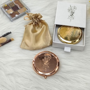 Puede incluir: Un espejo compacto de oro rosa con grabado floral y el nombre "Erisay". También se ve una bolsa dorada de satén con cordón, una caja blanca con un espejo compacto dorado en el interior y una paleta de maquillaje. Los artículos están sobre una superficie blanca y esponjosa.