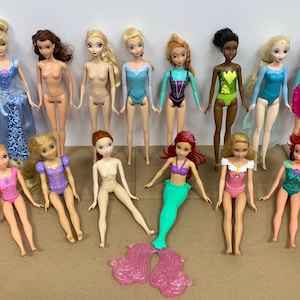 K&ouml;nnte beinhalten: Eine Sammlung von Modepuppen, darunter Prinzessinnen und Meerjungfrauen. Die Puppen haben verschiedene Haarfarben und tragen Badeanz&uuml;ge oder Kleider in Blau-, Rosa-, Gr&uuml;n- und Lilat&ouml;nen. Einige Puppen sind nackt.