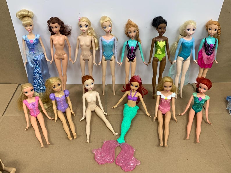 K&ouml;nnte beinhalten: Eine Sammlung von Modepuppen, darunter Prinzessinnen und Meerjungfrauen. Die Puppen haben verschiedene Haarfarben und tragen Badeanz&uuml;ge oder Kleider in Blau-, Rosa-, Gr&uuml;n- und Lilat&ouml;nen. Einige Puppen sind nackt.