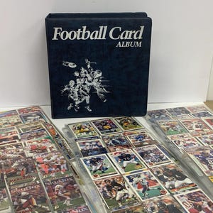 May include: A navy blue football card album with the words "Football Card ALBUM" in white. The album is next to several plastic sheets filled with football trading cards. The cards feature various players in action.