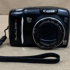 May include: A black Canon PowerShot SX110 IS digital camera with a 10x optical zoom lens. The front features the text Canon, PowerShot SX110 IS, 10x Optical Zoom, 9.0 Mega Pixels, and lens specifications 6.0-60.0mm 1:2.8-4.3. Includes a black wrist strap.