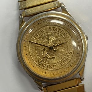 May include: A gold-tone wristwatch featuring a textured gold dial with the United States Marine Corps emblem, including an eagle, globe, and anchor. The watch has a matching gold-tone expansion band and a round case with a silver-tone bezel.