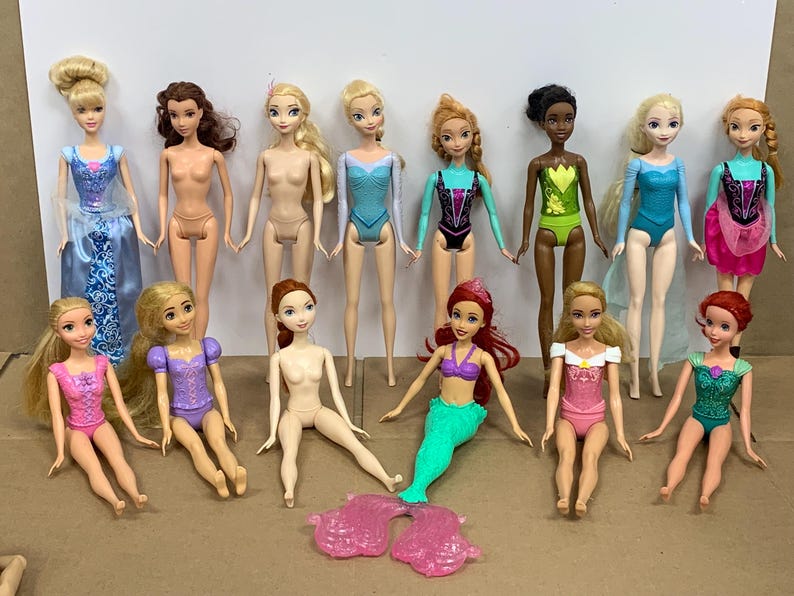 K&ouml;nnte beinhalten: Eine Sammlung von Modepuppen mit verschiedenen Hautt&ouml;nen und Frisuren. Einige Puppen tragen bunte Badeanz&uuml;ge oder Kleider, andere sind unbekleidet. Eine Puppe hat eine Meerjungfrauenflosse. Die Puppen sind auf einer neutralen Oberfl&auml;che angeordnet.