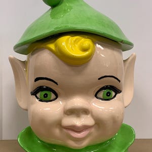 May include: A ceramic cookie jar shaped like a smiling elf's head. The jar features a green hat, collar, and eyes, with yellow hair and a pale pink face. The elf has large green eyes and pointed ears. The jar is a vintage collectible.