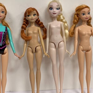 May include: Four plastic dolls standing in a row. The first doll on the left wears a turquoise and black leotard. The other three dolls are unclad, featuring molded skin-toned undergarments. Two dolls have auburn hair in braids, one has blonde hair in a side braid, and one has long auburn hair.
