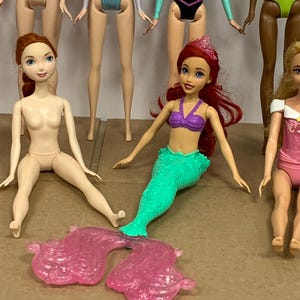 K&ouml;nnte beinhalten: Eine Sammlung von Modepuppen, darunter eine Meerjungfrauenpuppe mit gr&uuml;nem Schwanz und lila Oberteil sowie eine Puppe mit rotem Haar. Weitere Puppen sind im Hintergrund zu sehen, mit verschiedenen Outfits.