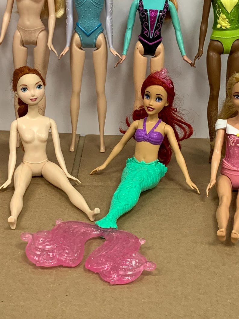 K&ouml;nnte beinhalten: Eine Sammlung von Modepuppen, darunter eine Meerjungfrauenpuppe mit gr&uuml;nem Schwanz und lila Oberteil sowie eine Puppe mit rotem Haar. Weitere Puppen sind im Hintergrund zu sehen, mit verschiedenen Outfits.