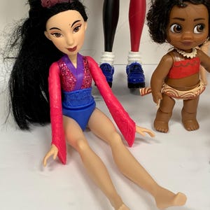 May include: A doll with long black hair and a purple crown, wearing a pink glittery top with long sleeves and a blue bottom. Beside it stands a smaller doll with curly dark hair, wearing a red top and a tan skirt with a white shell necklace.