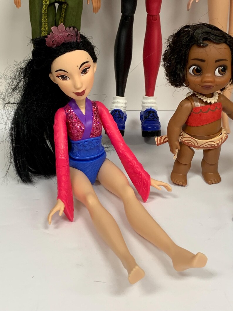 May include: A doll with long black hair and a purple crown, wearing a pink glittery top with long sleeves and a blue bottom. Beside it stands a smaller doll with curly dark hair, wearing a red top and a tan skirt with a white shell necklace.