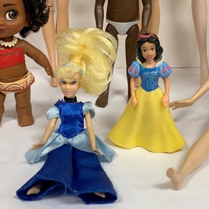 May include: A collection of small dolls including a figure with dark curly hair in a red and tan outfit, a blonde doll in a blue satin dress, a dark-skinned doll in white briefs, a Snow White figure in a blue and yellow dress, and a doll in a blue top.