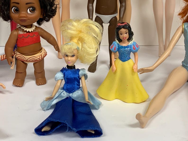 May include: A collection of small dolls including a figure with dark curly hair in a red and tan outfit, a blonde doll in a blue satin dress, a dark-skinned doll in white briefs, a Snow White figure in a blue and yellow dress, and a doll in a blue top.