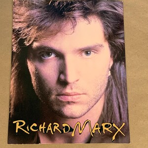 May include: A vintage promotional photo featuring a man with long, wavy brown hair and a serious expression. The text "Richard Marx" is written in a stylized font across the bottom of the image.
