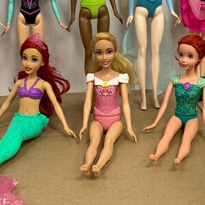K&ouml;nnte beinhalten: Mehrere Modepuppen werden gezeigt, jede mit einzigartigen Outfits und Frisuren. Eine Puppe hat rotes Haar und ein lila Oberteil mit einem gr&uuml;nen Meerjungfrauenschwanz. Eine andere Puppe tr&auml;gt einen rosa Badeanzug mit einem wei&szlig;en Oberteil. Die Puppen befinden sich auf einer braunen Oberfl&auml;che.