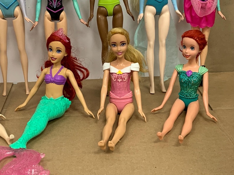 K&ouml;nnte beinhalten: Mehrere Modepuppen werden gezeigt, jede mit einzigartigen Outfits und Frisuren. Eine Puppe hat rotes Haar und ein lila Oberteil mit einem gr&uuml;nen Meerjungfrauenschwanz. Eine andere Puppe tr&auml;gt einen rosa Badeanzug mit einem wei&szlig;en Oberteil. Die Puppen befinden sich auf einer braunen Oberfl&auml;che.