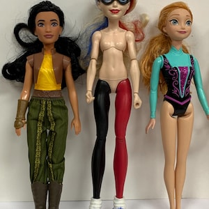 May include: Three dolls standing side by side. The left doll has long black hair, a yellow top, brown vest, green pants, and brown boots. The center doll has blonde hair with blue and red streaks, a black mask, and black and red leggings. The right doll has orange hair and a teal and black leotard.