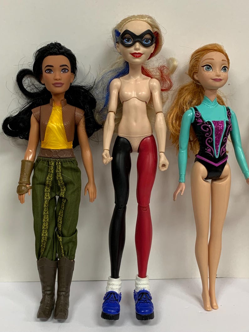 May include: Three dolls standing side by side. The left doll has long black hair, a yellow top, brown vest, green pants, and brown boots. The center doll has blonde hair with blue and red streaks, a black mask, and black and red leggings. The right doll has orange hair and a teal and black leotard.