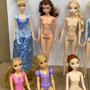 K&ouml;nnte beinhalten: Eine Sammlung von Modepuppen mit verschiedenen Charakteren. Eine Puppe tr&auml;gt ein blaues Kleid mit silbernem Oberteil und weitem Rock. Andere Puppen haben unterschiedliche Haarfarben und Outfits, darunter rosa, lila und blond.