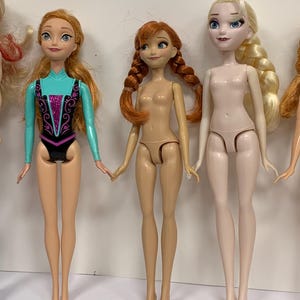 May include: Three plastic dolls standing in a row. The left doll has long orange hair and wears a turquoise and black leotard with purple glitter. The middle doll has orange hair in two braids. The right doll has long blonde hair in a side braid.