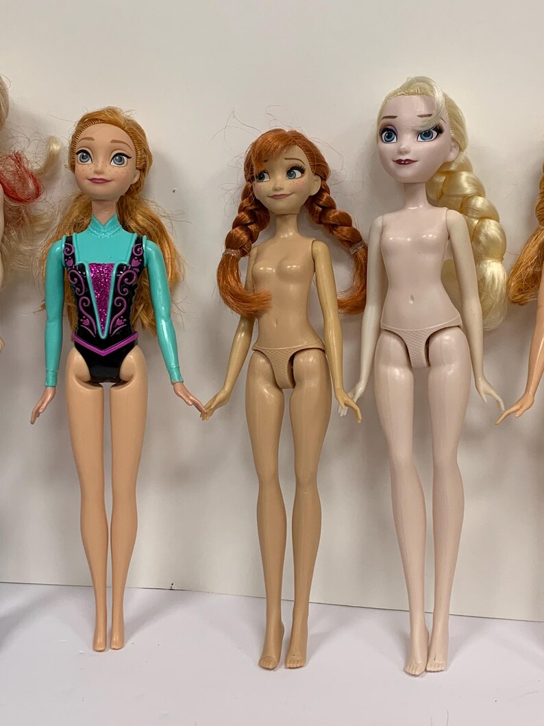 May include: Three plastic dolls standing in a row. The left doll has long orange hair and wears a turquoise and black leotard with purple glitter. The middle doll has orange hair in two braids. The right doll has long blonde hair in a side braid.
