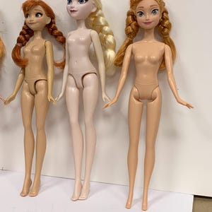 May include: Three plastic dolls standing in a row. The left doll has auburn hair in two braids. The center doll has platinum blonde hair in a side braid and pale skin. The right doll has light brown hair in two braids. All dolls are unclothed.