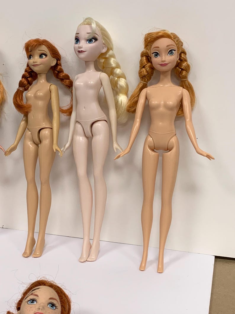 May include: Three plastic dolls standing in a row. The left doll has auburn hair in two braids. The center doll has platinum blonde hair in a side braid and pale skin. The right doll has light brown hair in two braids. All dolls are unclothed.