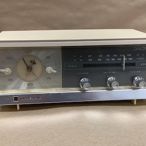 May include: A vintage Panasonic solid state clock radio with a cream-colored rectangular casing. The front features an analog clock face on the left and a dark gray radio tuner panel on the right with FM and AM frequency scales and three knobs.