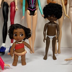 May include: A collection of various dolls including a small figure with dark curly hair wearing a red top and patterned skirt, and a dark-skinned doll with short curly hair wearing white underwear, surrounded by several other partially visible dolls.