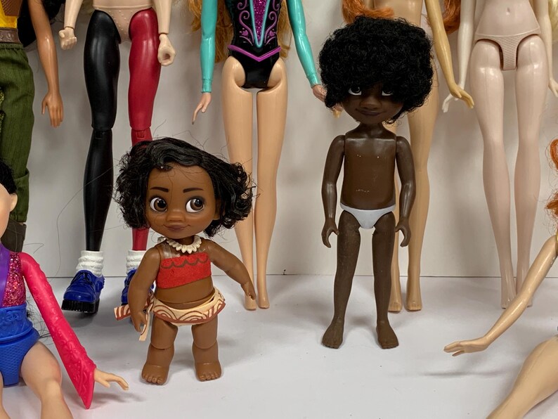 May include: A collection of various dolls including a small figure with dark curly hair wearing a red top and patterned skirt, and a dark-skinned doll with short curly hair wearing white underwear, surrounded by several other partially visible dolls.