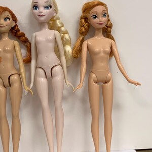 May include: Three plastic dolls standing side by side. The left doll has auburn hair in braids. The center doll has platinum blonde hair in a side braid and pale skin. The right doll has strawberry blonde hair in two braids and tan skin.