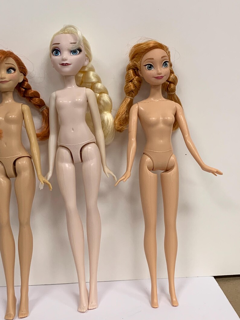 May include: Three plastic dolls standing side by side. The left doll has auburn hair in braids. The center doll has platinum blonde hair in a side braid and pale skin. The right doll has strawberry blonde hair in two braids and tan skin.