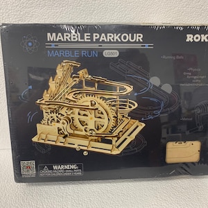May include: A black rectangular box for a ROKR Marble Parkour marble run model kit, model LG501. The box features a graphic of the assembled wooden gear-driven marble track, along with text labels for running balls, physics, and manual operation.