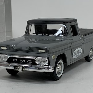 May include: A gray 1960 GMC pickup truck model with a black roof and white-walled tires. The truck has chrome detailing on the front bumper and grill. The text "1960 GMC" is visible on the license plate. The side of the truck has a white oval logo with text.