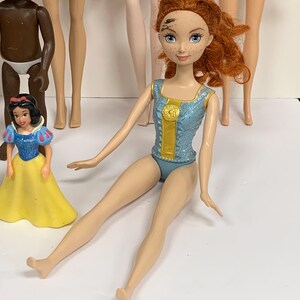 May include: A doll with curly red hair, blue eyes, and freckles, wearing a light blue glittery leotard with a gold center panel. The doll is seated, surrounded by other dolls, including a small Snow White figure and a dark-skinned doll.