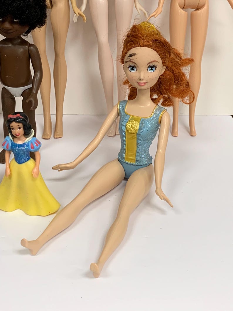 May include: A doll with curly red hair, blue eyes, and freckles, wearing a light blue glittery leotard with a gold center panel. The doll is seated, surrounded by other dolls, including a small Snow White figure and a dark-skinned doll.