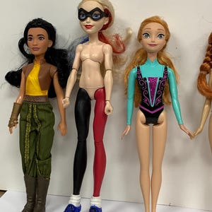 May include: Three dolls standing side by side. The first has black hair, a yellow top, brown vest, and green pants. The second has blonde hair with red and blue streaks, a black mask, and black and red leggings. The third has orange hair and a teal leotard.