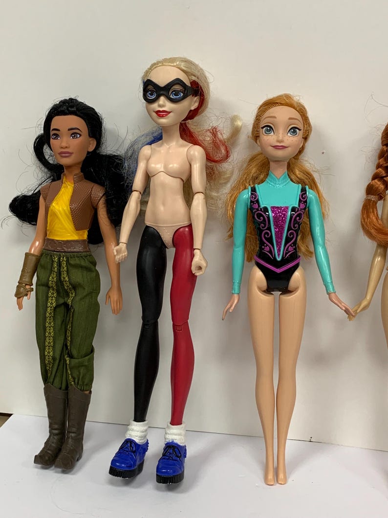 May include: Three dolls standing side by side. The first has black hair, a yellow top, brown vest, and green pants. The second has blonde hair with red and blue streaks, a black mask, and black and red leggings. The third has orange hair and a teal leotard.