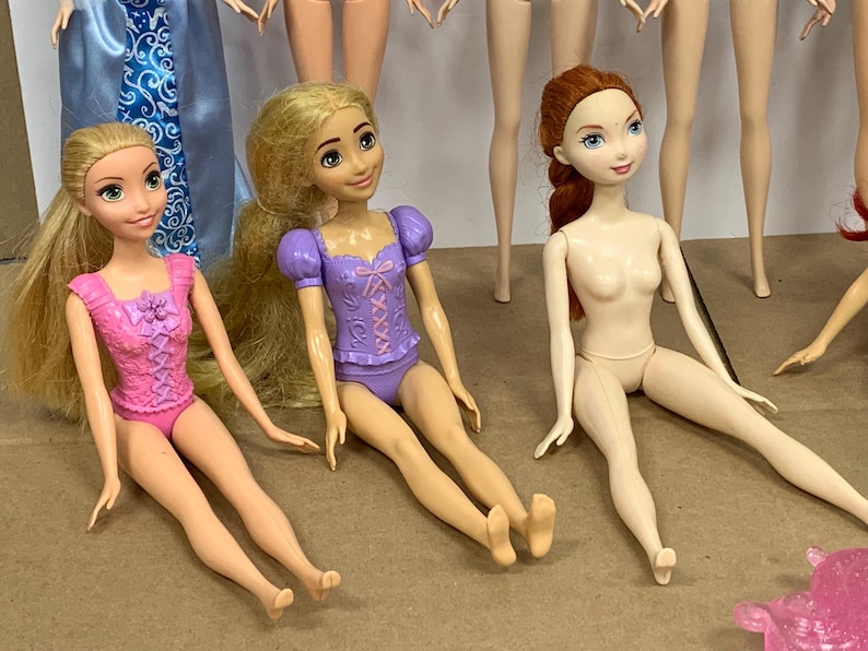 K&ouml;nnte beinhalten: Mehrere Modepuppen werden gezeigt, jede mit einzigartigen Merkmalen. Zwei Puppen haben lange blonde Haare und tragen rosa und lila Outfits. Eine weitere Puppe mit roten Haaren ist unbekleidet. Die Puppen sitzen.