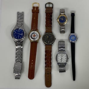 May include: A collection of six wristwatches with various designs. The watches feature metal and leather bands, with faces in blue, white, and gold. Some have analog displays, while others have digital displays. The watches are arranged on a white surface.