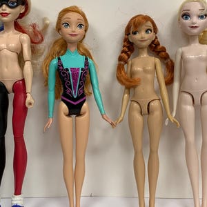 May include: Four dolls standing in a row. The first doll has blonde hair, a black mask, and red and black leggings. The second doll has long orange hair and a turquoise and black leotard. The third doll has orange braided hair. The fourth doll has blonde hair.