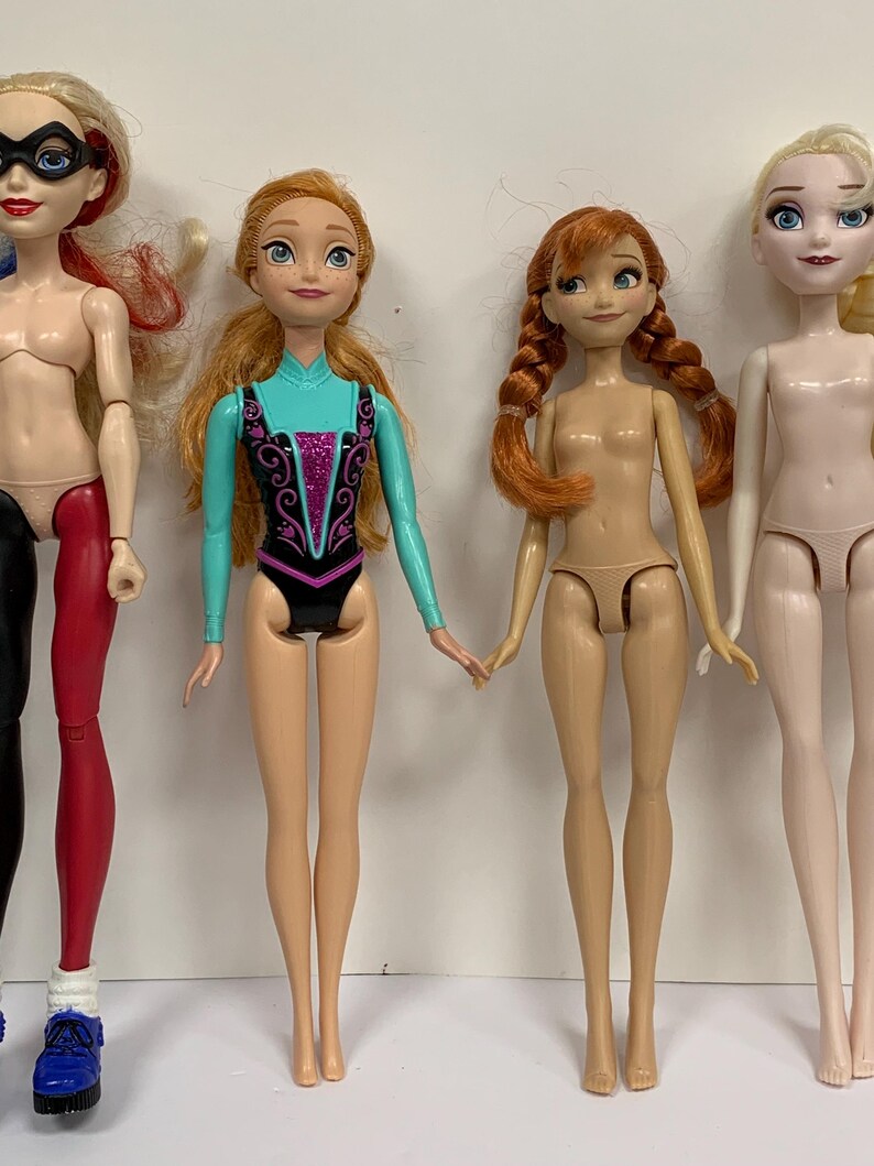 May include: Four dolls standing in a row. The first doll has blonde hair, a black mask, and red and black leggings. The second doll has long orange hair and a turquoise and black leotard. The third doll has orange braided hair. The fourth doll has blonde hair.