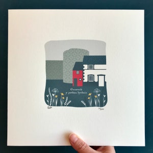 May include: A colourful illustration of a red house with a grey roof and a green background. The house is surrounded by yellow flowers and green grass. The text "Gwynnech y pethau bychain" is written below the house.