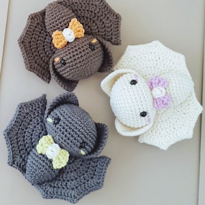 May include: Three crocheted stuffed animals shaped like bats. One bat is brown with orange and white accents, one is gray with yellow accents, and one is white with pink accents.