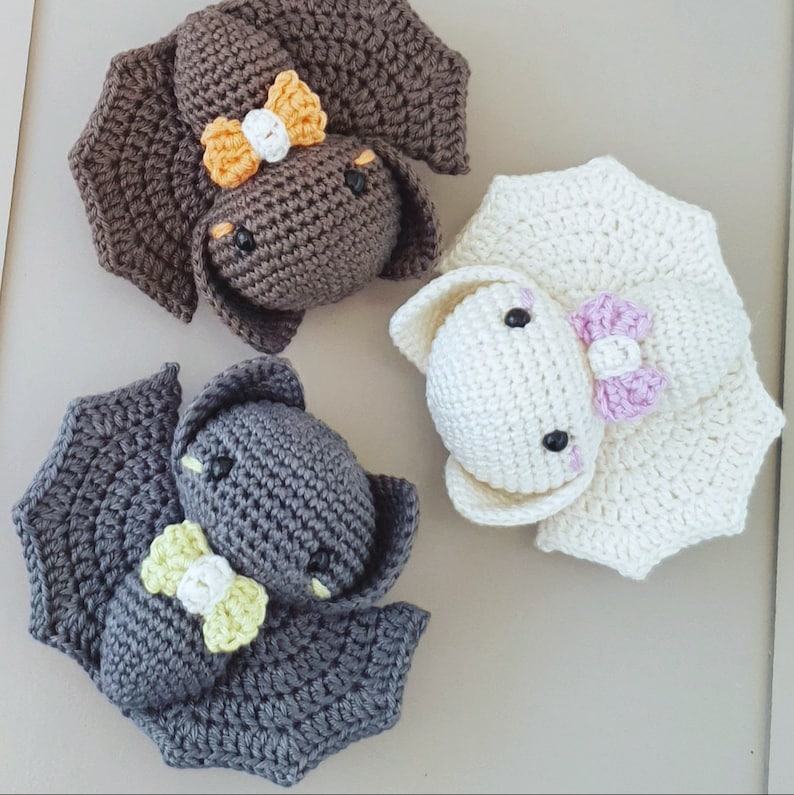 May include: Three crocheted stuffed animals shaped like bats. One bat is brown with orange and white accents, one is gray with yellow accents, and one is white with pink accents.